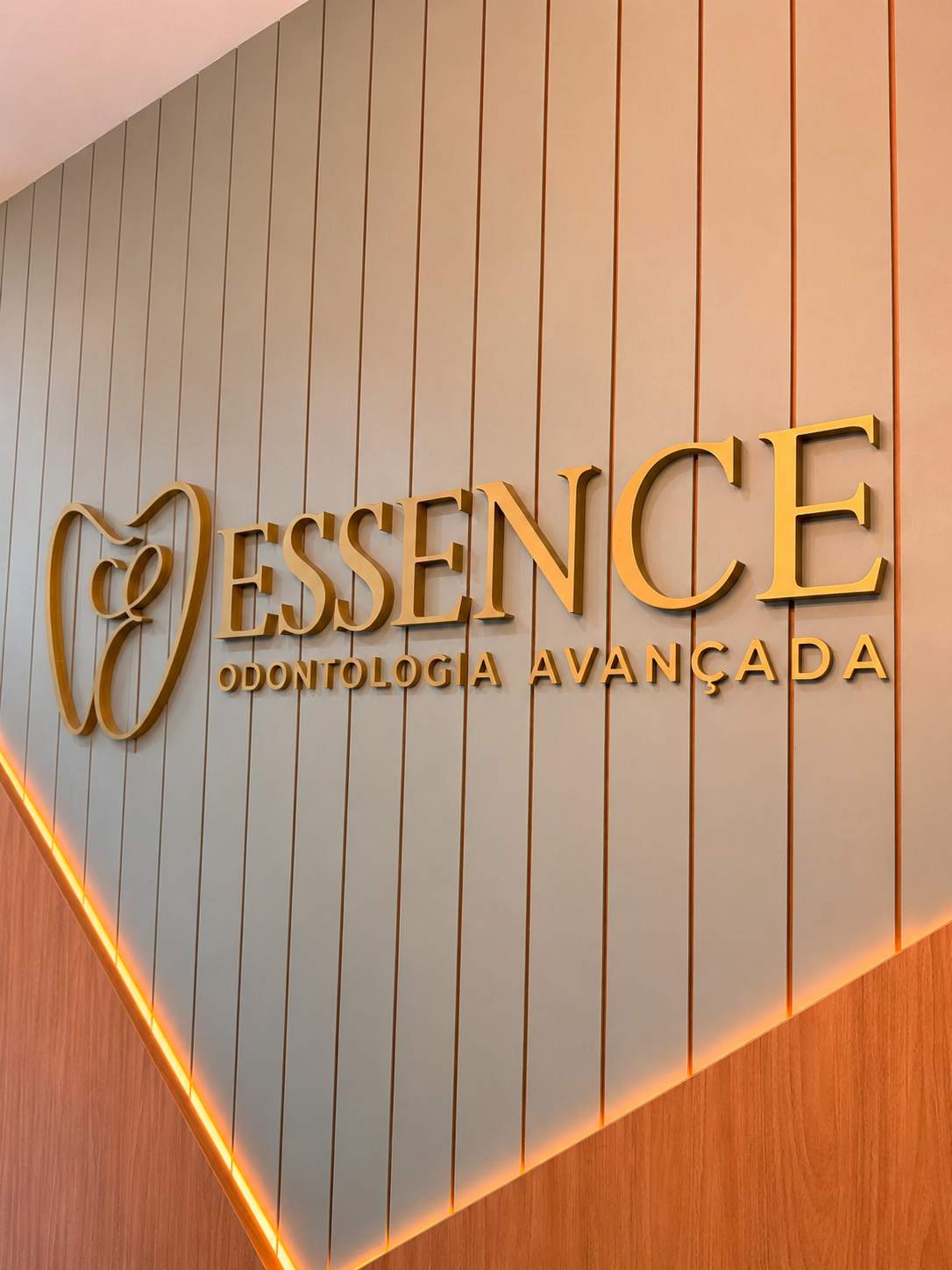 Parede da recepção da Essence com a marca em letras douradas sobre ripado verde e um rasgo de luz âmbar na diagonal
